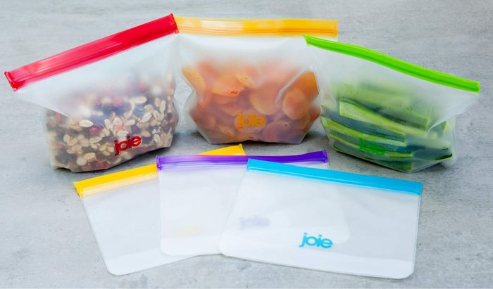 Joie brand reusable storage bags, some containing nuts, dried fruit, and cucumber slices, are showcased on a surface, emphasizing their adaptability for storing various foods.
