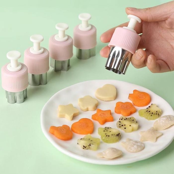 A hand using a pink and silver vegetable cutter to shape fruits and vegetables for a white plate, with four similar cutters visible in the background
