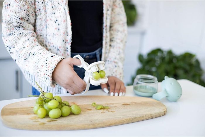 An individual using a grape cutter to slice green grapes on a wooden cutting board, with a whale-shaped accessory nearby.
