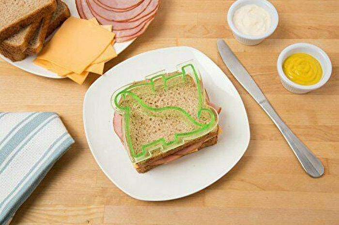 A sandwich on a plate with a green dinosaur-shaped cutter on top, accompanied by ham, cheese, mayo, mustard, and a knife