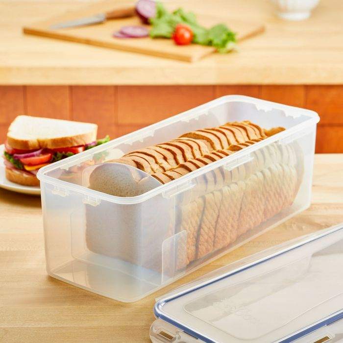 Sliced bread kept in a transparent plastic container on a kitchen counter, with a sandwich on a plate and chopped vegetables on a cutting board in the background.
