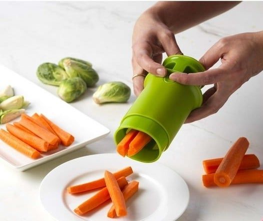 A hand uses a green cylindrical kitchen gadget to chop carrots onto a plate.