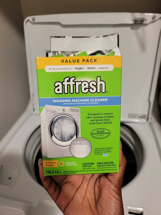 A hand holds a box of Affresh washing machine cleaner tablets in front of an open washing machine, showing an image of a washing machine along with product details on the box.