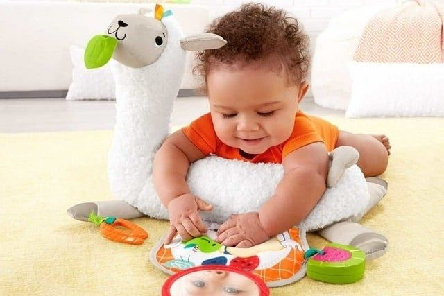 An infant sitting comfortably on a plush tummy wedge toy, which features various textures and colorful tags, designed for babies as young as 0+ months.