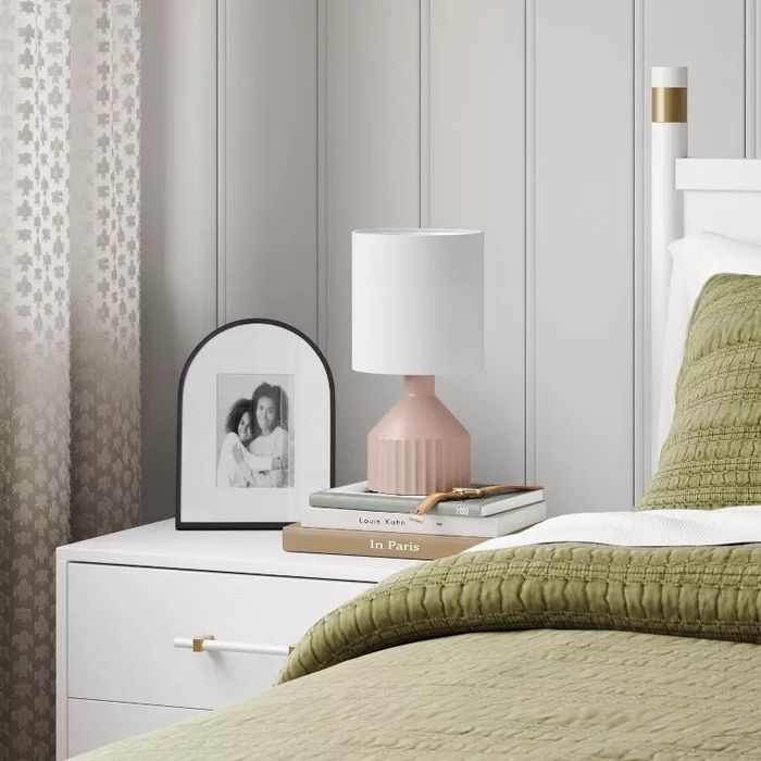 A neatly made bed with a green quilt, beside a white nightstand with a pink lamp, books, and a framed black-and-white photo, in a cozy bedroom