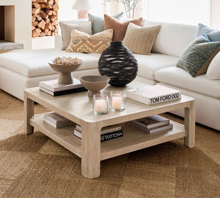 A modern living room with a white sectional sofa, a wooden coffee table adorned with decorative bowls, vases, candles, and books, including 'Tom Ford 002.'