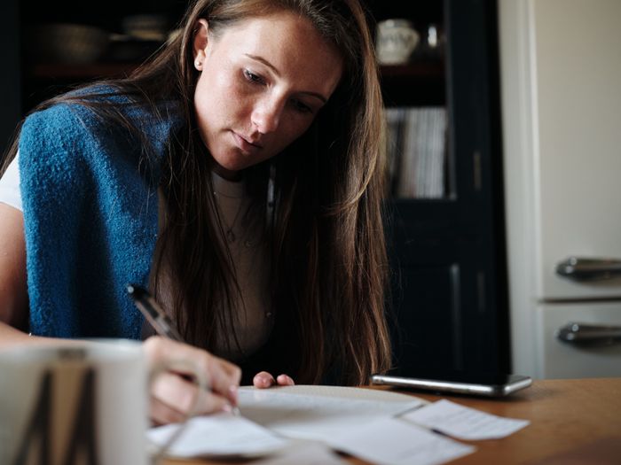 A person takes notes at a table, a cup resting beside them and a phone nearby. A soft blue blanket drapes over their shoulders, giving the scene a cozy and reflective vibe.