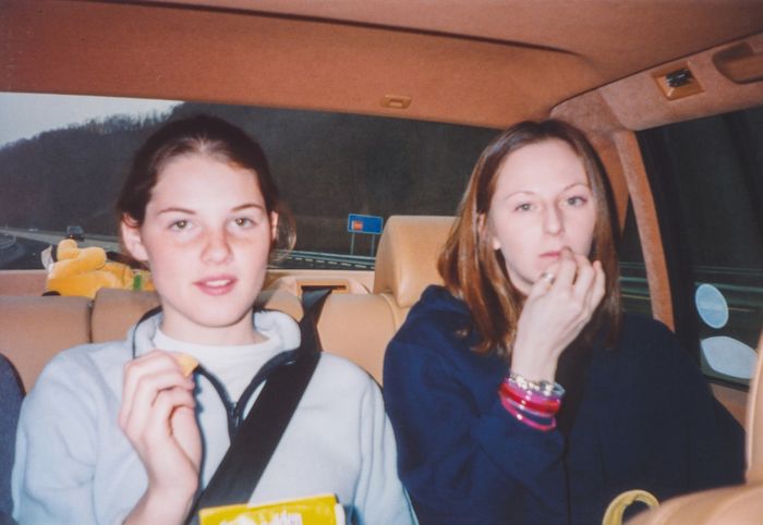 Two people sit in a car, enjoying snacks and looking at the camera, with a plush toy sitting on the seat in the background.