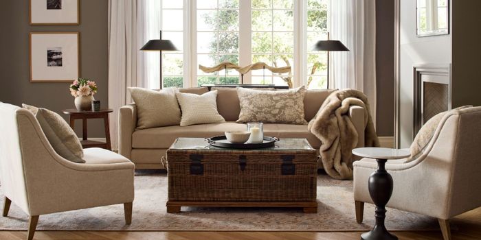 A cozy living room featuring a beige sofa, two cushioned chairs, a wicker trunk used as a coffee table, and large windows letting in ample natural light.