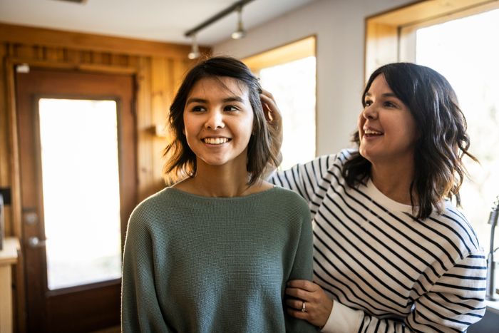 Two people share a warm moment indoors, with one adjusting the other's hair. Both are smiling in a cozy environment adorned with wooden elements.