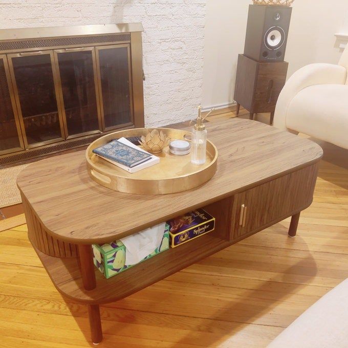 A wooden coffee table with storage shelves, displaying books, a gold tray with decorative items, and a notebook, placed in a cozy living room.