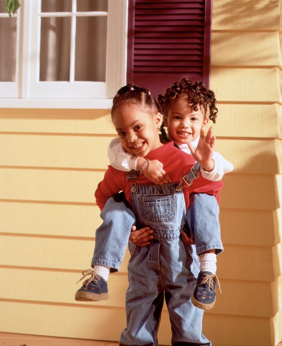 Two joyful children play outside, one giving the other a piggyback ride. They both wear comfortable, casual overalls and sweaters.