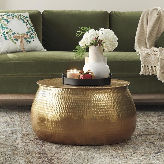 A gold hammered metal coffee table adorned with candles, flowers, and gifts, set against a green sofa with decorative pillows and a cozy throw blanket in the background.