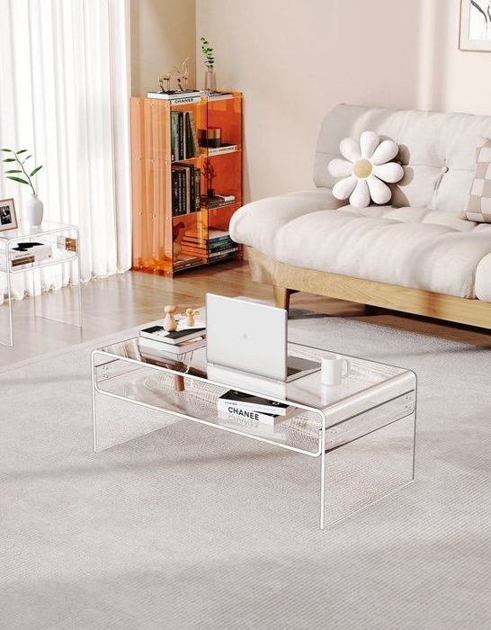 A modern living room featuring a clear coffee table with a laptop and a Chanel book, paired with an orange bookshelf and a beige couch with a daisy pillow.