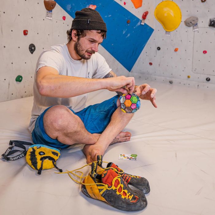 A person sitting on a gym floor holding a puzzle ball, with climbing shoes and a chalk bag nearby, in a bouldering area.