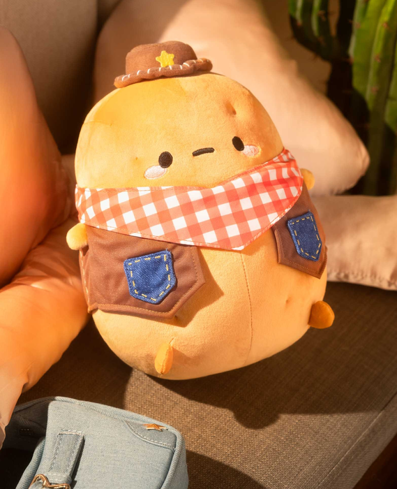 Plush toy resembling a potato dressed in a cowboy hat and a checkered bandana, styled as a cowboy, resting on a sofa with a cactus nearby