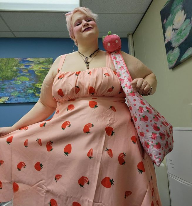 A person wearing a strawberry-patterned dress and a matching tote bag, smiling confidently indoors