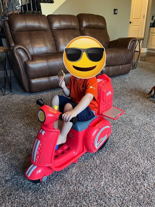 A child sitting on a toy scooter indoors, wearing a shirt and holding something in their hand.