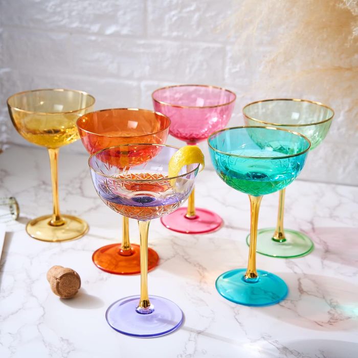 Six elegant cocktail glasses with gold stems and translucent, colorful bowls, arranged on a marble surface with a cork nearby.