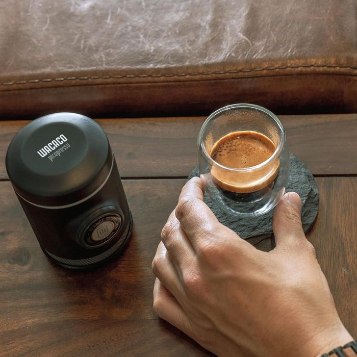 A hand holds a glass of espresso next to a Wacaco Nanopresso portable coffee maker on a wooden surface