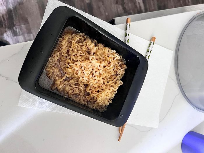 A bowl of cooked instant ramen noodles resting on a white paper napkin, with chopsticks beside it. A portion of a gray surface is visible in the corner