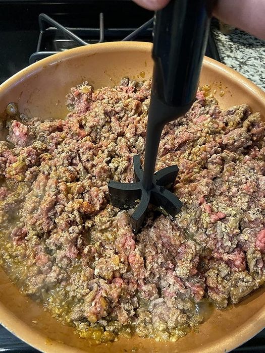 Ground beef sizzles in a skillet as a hand uses a meat chopper tool to break it up