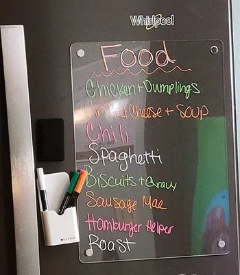A clear rectangular dry-erase board hung vertically on a fridge, displaying a list of ingredients