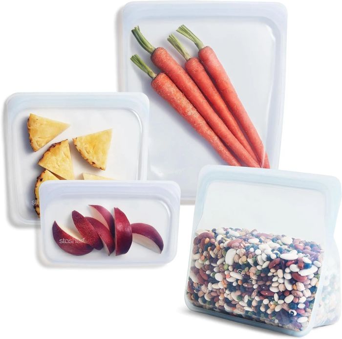 Four reusable silicone Stasher bags filled with different produce: one with carrots, one with pineapple slices, one with apple slices, and one with mixed beans