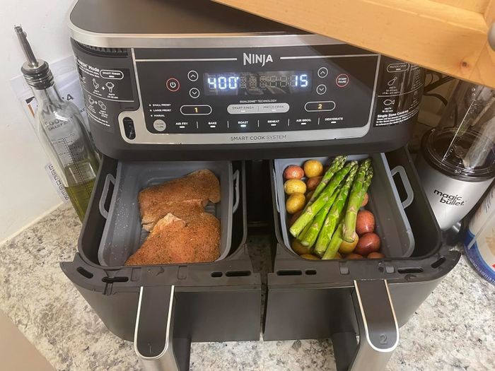 A Ninja air fryer with two compartments: one holding seasoned meat and the other with asparagus and small potatoes. A Magic Bullet blender sits on the right
