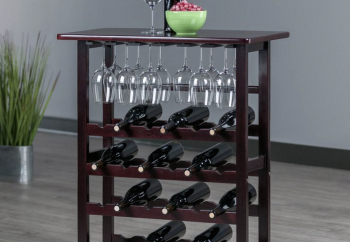 A wooden wine rack elegantly holding 11 bottles of wine in a vertical position, with wine glasses hanging beneath the tabletop. A wine bottle, wine glass, and a bowl of grapes rest on top of the rack.