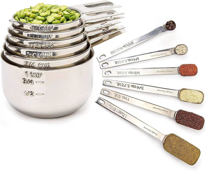 A set of metal measuring cups filled with green split peas, accompanied by measuring spoons with spices. Numbers and measurements are clearly visible on each item.