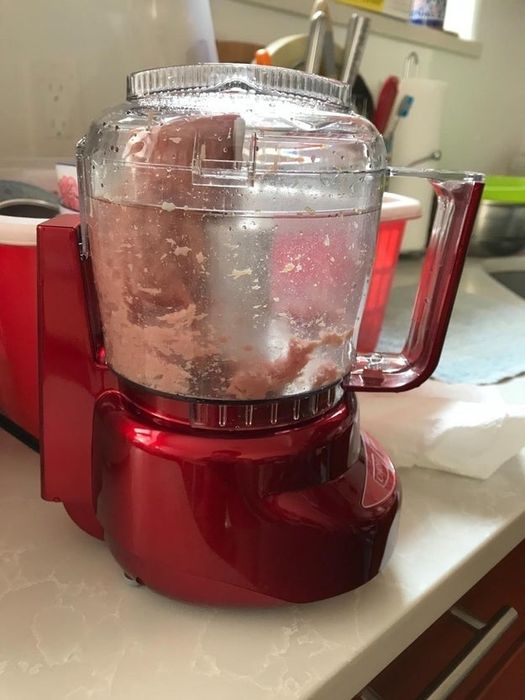 A food processor on a kitchen counter with a mix of chopped pink meat inside. Various kitchen tools are visible in the background