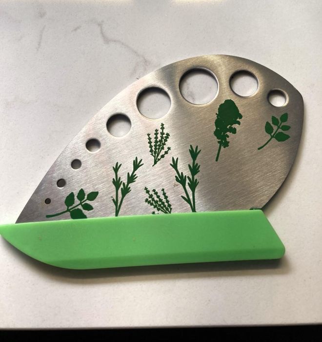 Metal herb stripper tool featuring a green protective guard placed on a white surface. The tool showcases herb illustrations and includes various sized holes for stripping leaves