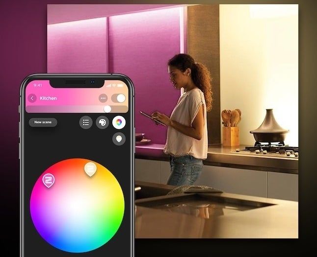 A woman in the kitchen controlling smart lighting with her phone, adjusting it to 16 million colors. The phone screen shows the lighting control app.