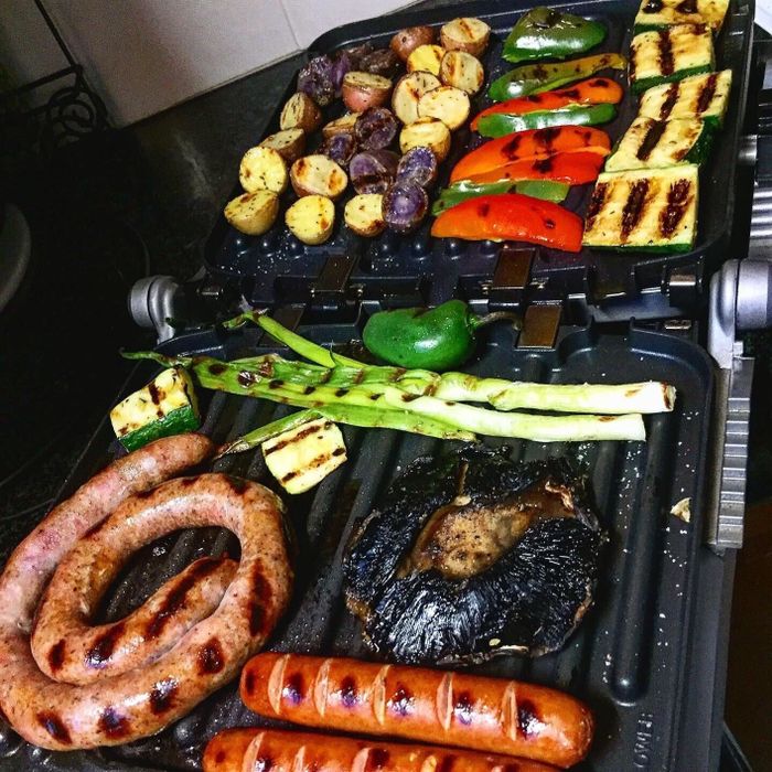 A variety of grilled foods on a grill, including sausages, hot dogs, mushrooms, asparagus, zucchini, bell peppers, and different types of potatoes