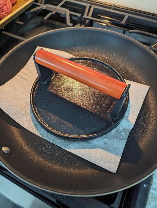 A round, cast-iron grill press with a red handle, branded 'Chefgrill,' resting on parchment paper inside a frying pan on the stovetop