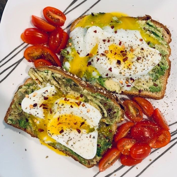 Sliced avocado toast topped with poached eggs, sprinkled with red chili flakes, accompanied by halved cherry tomatoes on a white plate