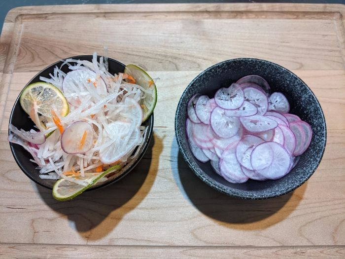 Two black bowls on a wooden surface: one filled with a mixed vegetable salad featuring sliced radish, lemon, and other veggies, and the other containing thinly sliced radishes