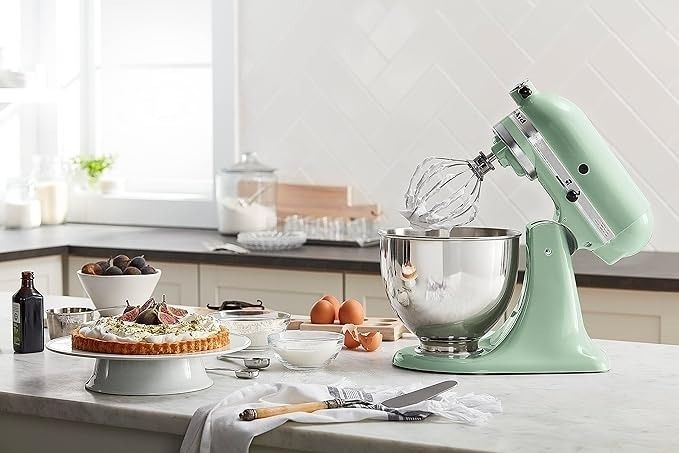 A kitchen counter full of tools, including a stand mixer, a cake topped with fruits on a cake stand, along with various baking ingredients and utensils.