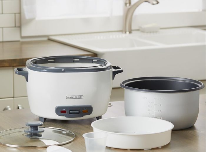 A Black+Decker rice cooker, complete with cooking base, bowl, steaming basket, glass lid, serving spoon, and a rice measuring cup placed on a kitchen counter