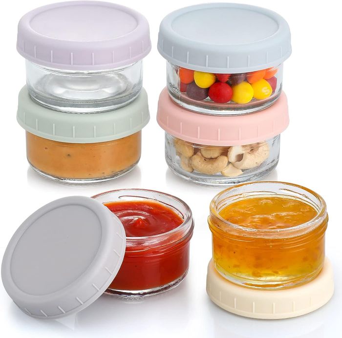 Four stackable glass jars with different contents, such as spreads and snacks, each featuring pastel-colored lids