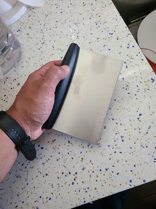 A person's hand grips an OXO brand dough scraper over a speckled countertop