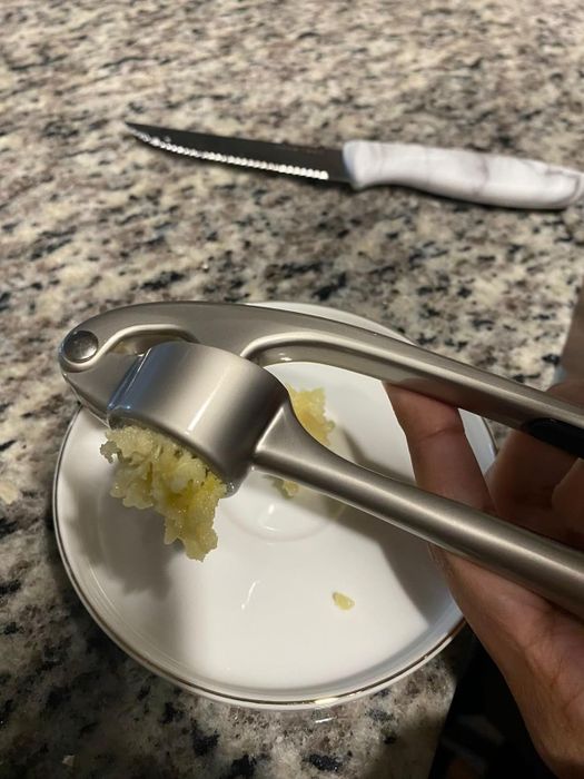A hand pressing garlic over a white plate on a granite countertop, with a serrated knife with a white handle in the background