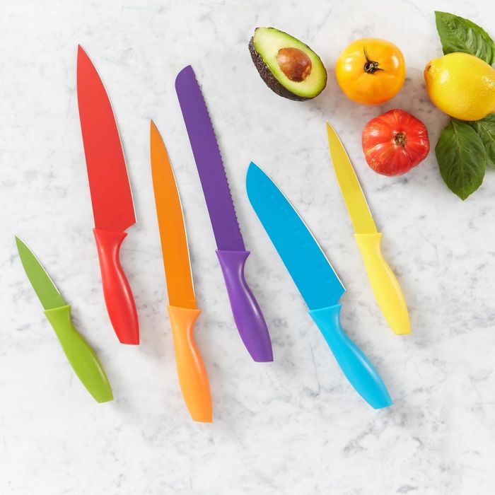 A lively kitchen knife set consisting of seven knives, displayed alongside an assortment of vegetables and fruits including avocado, lemon, orange, and tomato, all arranged on a polished marble surface.