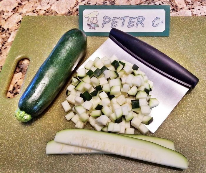A cutting board holding chopped and whole zucchini, half-sliced zucchini, a chopper tool, and a sign reading 