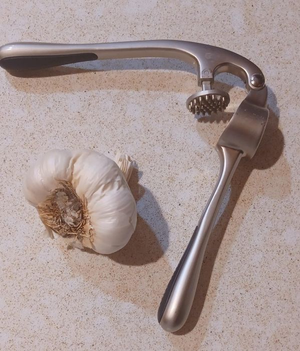 A metallic garlic press placed alongside a whole garlic bulb on a speckled countertop