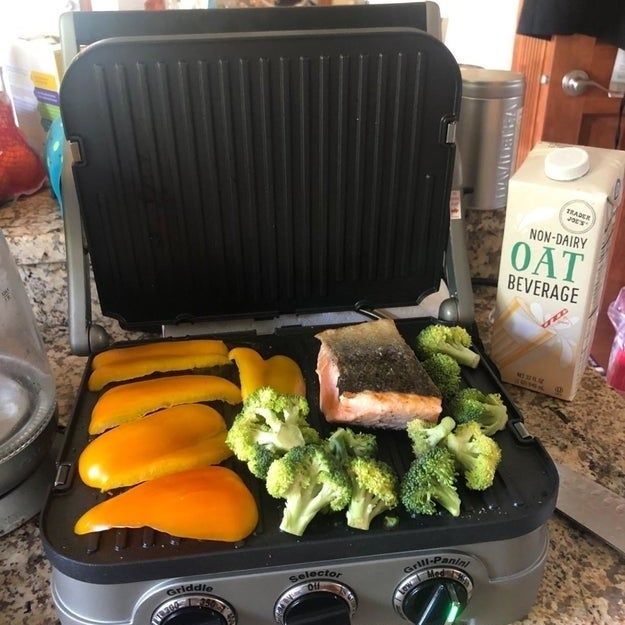 A kitchen counter featuring a stovetop grill cooking yellow bell peppers, broccoli, and salmon. A carton of non-dairy oat beverage rests beside the grill