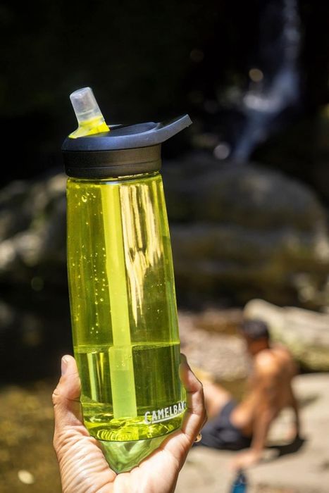 Person holding a CamelBak water bottle with another individual blurred in the background near water