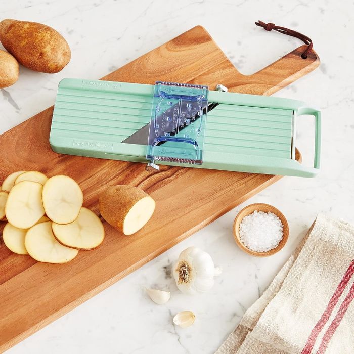 A mandoline slicer on a wooden cutting board, accompanied by sliced potatoes, a whole potato, garlic cloves, sea salt, and a striped cloth nearby