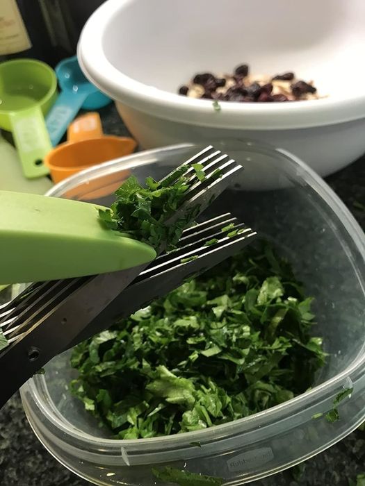 Scissors cutting herbs over a plastic container, with a bowl and measuring spoons in the background